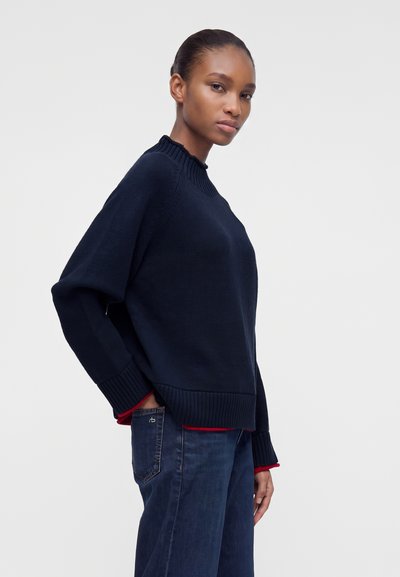Jeune personne portant un pull bleu marine et un jean bleu, debout avec une main dans la poche, regardant la caméra sur un fond uni.