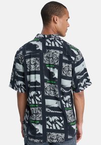 Κοντομάνικο πουκάμισο σε ναυτικό μπλε με ασπρόμαυρα γραφικά σχέδια. Διαθέτει διάφορα κυκλικά και μοτίβα σανίδας σερφ, με πράσινες επισημάνσεις κειμένου.
