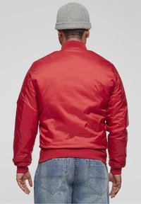 Giacca bomber rossa con una texture liscia e lucida e polsini a costine, abbinata a jeans di denim blu e un berretto grigio di maglia.
