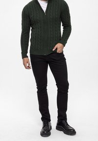 Groen kabelgebreid vest met ritssluiting aan de voorkant, geribde manchetten en zoom. Gecombineerd met nauwsluitende zwarte jeans en zwarte veterschoenen.