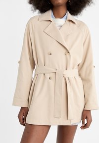 Beige dubbelgeknoopte trenchcoat met riem, gemaakt van een zacht materiaal. Heeft schouderknopen en een losse pasvorm, lengte boven de knie.