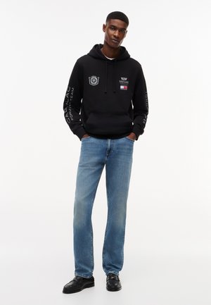 Jauns vīrietis stāv ar rokām kabatās, valkājot melnu Cadillac Formula 1 džemperi ar kapuci, zilas džinsas un melnas loafer kurpes, baltā fonā.