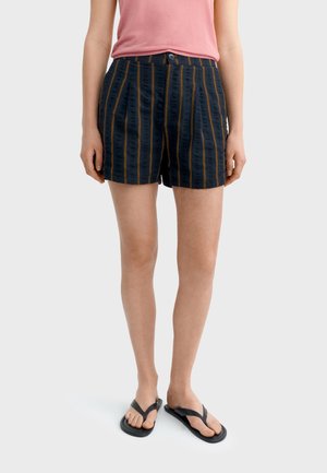 Frau trägt schwarze Shorts mit vertikalen orangen Streifen, ein pinkfarbenes ärmelloses Oberteil und schwarze Flip-Flops, steht vor hellem Hintergrund.