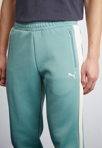 Teal Sporthosen aus strukturiertem Material, mit einem weißen seitlichen Streifen, elastischem Bund und einem kleinen weißen Puma-Logo am Oberschenkel.