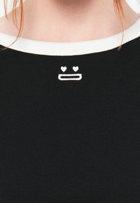 Svart ribbestrikket topp med hvit rund hals som har et enkelt broderi av et ansikt med hjerter over en rett munn.