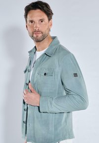 Hellgrüne Jacke aus weichem Stoff, mit knöpfbarem Vorderteil, zwei Brusttaschen und einem umgeschlagenen Kragen. Glatte Textur mit dezentem Glanz.