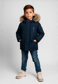 Boy standing with hands in pockets wearing navy parka with fur-lined hood, blue jeans, and beige sneakers against gray background.