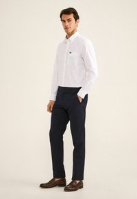 Camisa blanca de botones con un bolsillo azul claro, combinada con pantalones de un azul marino oscuro y mocasines marrones. Tejido suave y detalles de corte a medida.