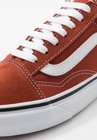 Röd mockaskor med vita snören och sidostrand, med svart gummisula och kontrasterande sömmar.