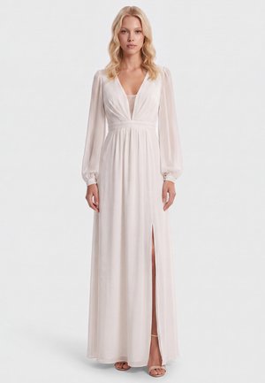Frau mit blondem Haar, die ein langes weißes Kleid mit transparenten langen Ärmeln und hohem Schlitz trägt, vor einem einfachen Hintergrund stehend.