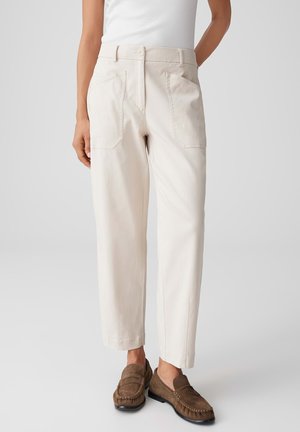 Vrouw draagt een hoge taille, recht model broek in off-white met voorzakken en bruine suède loafers tegen een effen achtergrond.