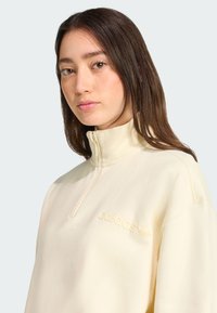 Sweatshirt zippé couleur crème avec col montant, ornée d'un logo Adidas brodé sur la poitrine et d'une texture lisse et douce.