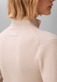 Suéter beige de cuello alto acanalado con cuello ajustado y detalles en las costuras laterales, con un pequeño logo blanco de Calvin Klein en la parte trasera.