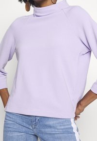 Frau, die einen ribbeligen, lavendelfarbenen Langarmoberteil mit hohem Kragen, blaue Jeans mit einem weißen Seitenstreifen und hölzernen Creolen trägt.