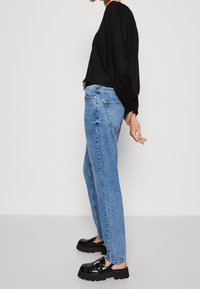 Person som bär en svart långärmad topp, ljusblå högmidjade jeans och svarta klumpiga loafers, stående på sidan mot en neutral bakgrund.