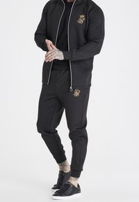 Giacca da pista nera con logo dorato, chiusura frontale con zip e pantaloni abbinati. Include polsini e tasche; abbinata a sneaker nere testurizzate.