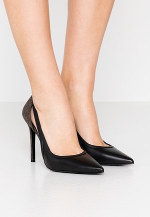 Czarne szpilki z wąskim, szpiczastym noskiem i cienkimi obcasami na bosej stopie na jednolitym białym tle.