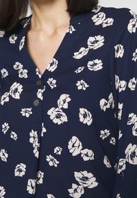 Detailní záběr na tmavě modrou blůzu s bílým květinovým potiskem a ozdobnými stříbrnými knoflíky u výstřihu a na manžetě rukávu.