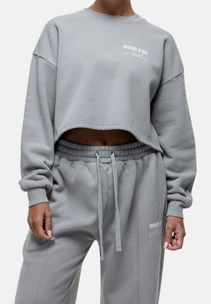 Femme portant un sweat-shirt court gris clair et un pantalon de jogging assorti avec le texte « Good For Nothing » sur les deux vêtements.