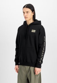 Sudadera negra de algodón con un bolsillo frontal, capucha con cordón y parche con logo en el pecho; presenta texto a lo largo de la manga derecha.