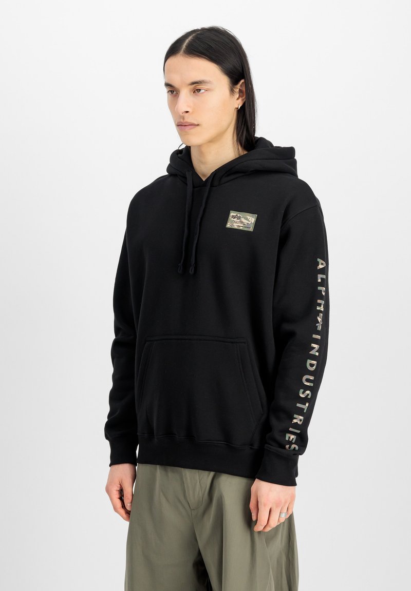 Sudadera negra de algodón con un bolsillo frontal, capucha con cordón y parche con logo en el pecho; presenta texto a lo largo de la manga derecha.
