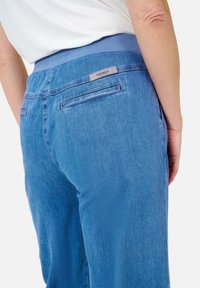 Personne portant un pantalon en denim bleu clair avec un waistband élastique et l'étiquette "Raphaela" au niveau de la poche arrière, une chemise blanche visible au-dessus du waistband.