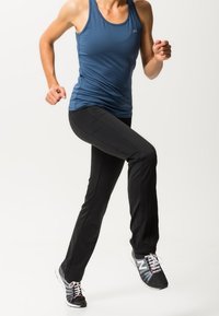 Osoba v modrém tílku a černých kalhotách běhá nebo se pohybuje na místě, má na sobě černobílé tenisky na bílém pozadí.