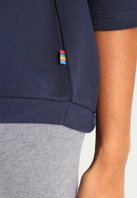 Top azul marino de manga corta con dobladillo acanalado y pequeña etiqueta de tela a rayas de colores, combinado con pantalones grises acanalados, mostrado en el brazo y la cintura de una persona.