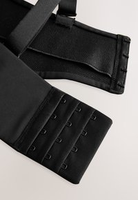 Gros plan d’une fermeture de vêtement en tissu noir avec des rangées de crochets et d’œillets ainsi que des bretelles réglables sur un fond clair.