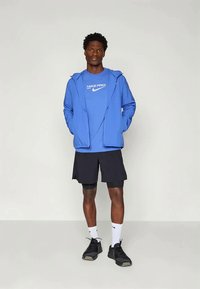 Hanorac Nike Pro albastru cu buzunare frontale, purtat peste un tricou albastru. Asortat cu pantaloni scurți negri și încălțăminte sport neagră. Sosete albe vizibile.