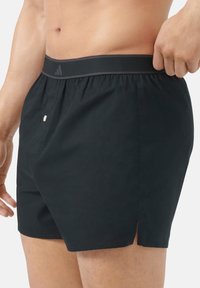 Černé plavecké šortky s šňůrkovým pasem, s bočními rozparky, hladkouTexturou a decentním logem podél pasu.