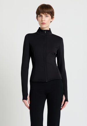 Donna che indossa una giacca nera aderente con cerniera, collo alto e fori per il pollice sulle maniche, abbinata a pantaloni neri, in piedi su sfondo bianco.