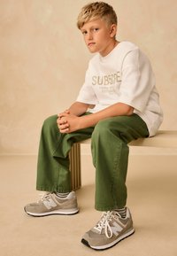 Šedé tenisky New Balance s bílými tkaničkami, zelené kalhoty a bílá košile s potiskem. Hladká látka a strukturovaný design.