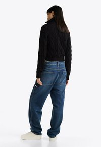 Maglione a collo alto in maglia lavorata a trecce nero abbinato a jeans blu dal taglio ampio. I jeans presentano cuciture a contrasto e tasche posteriori. Scarpe da ginnastica bianche.