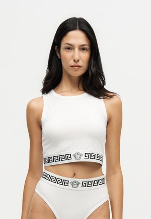 Donna con lunghi capelli scuri indossa un top corto bianco senza maniche e slip a vita alta abbinati con fasce nere con motivo a chiave greca e testa di Medusa.
