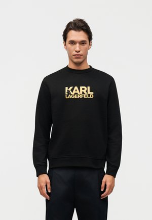 Uomo con capelli scuri che indossa una felpa nera con il logo dorato "KARL LAGERFELD", in piedi con le mani rilassate contro uno sfondo grigio chiaro.