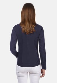 Navyblaues Langarmhemd mit glatter, glänzender Oberfläche, ausgestattet mit einer taillierten Passform und Knopfmanschetten, von hinten mit weißen Hosen gezeigt.