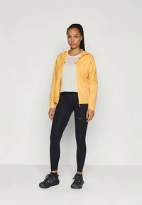 Veste légère jaune avec une capuche, superposée à une chemise blanche, associée à des leggings noirs et des chaussures de sport noires.