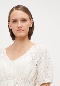 Weiße, strukturierte Bluse mit V-Ausschnitt, Puffärmeln und elastischem Bund. Sie verfügt über ein gewebtes Muster im gesamten Stoff.