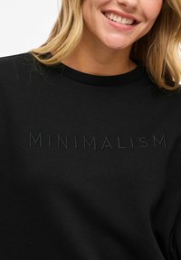 Smiling woman with blonde hair wears a black shirt embroidered with the word "MINIMALISM" across the chest.