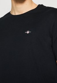 Maglietta nera in cotone con scollo rotondo, maniche corte e piccolo logo bianco e rosso sul petto in alto a sinistra. Tessuto morbido.