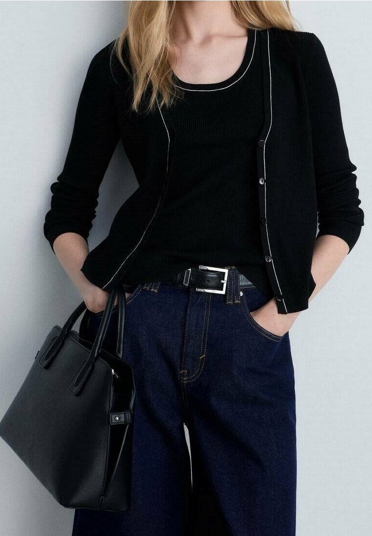 Femme portant un cardigan noir et un haut avec un jean bleu, tenant un sac à main noir, mains dans les poches, debout contre un mur uni.