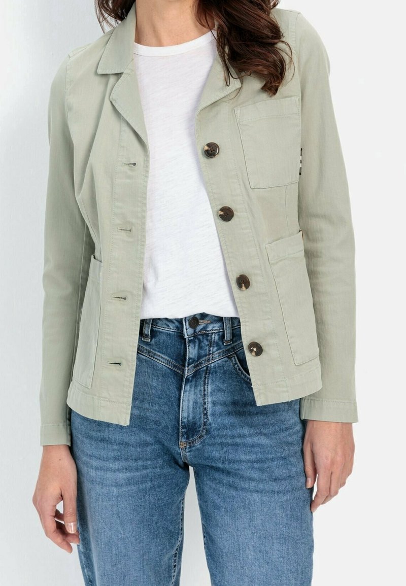 Femme portant une veste verte claire boutonnée sur un t-shirt blanc et un jean en denim bleu, debout devant un fond uni.