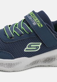 Mrežaste tenisice tamnoplave boje s zelenim naglascima, uz trakicu na čičak, teksturiranom bijelom potplatu i Skechers logom.