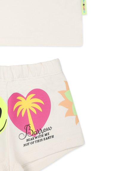 Jasno beżowa bluzka z krótkim rękawem połączona z wzorzystymi szortami, które mają na sobie uśmiechniętą buźkę, palmę, serce i słońce w jaskrawych kolorach.