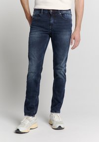 Donkerblauwe denim jeans met een slim fit, voorzien van voorzakken, lichte slijtage en subtiele stikdetails. Gecombineerd met witte sneakers.