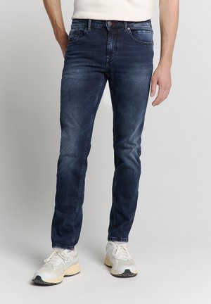 Dark blue denim jeans with a slim fit, featuring front pockets, slight fading, and subtle stitching details. Paired with white sneakers.