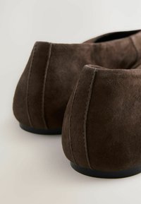 Le ballerine in suede marrone sono mostrate da dietro, mettendo in risalto i loro tacchi arrotondati e i dettagli della cucitura su uno sfondo chiaro.