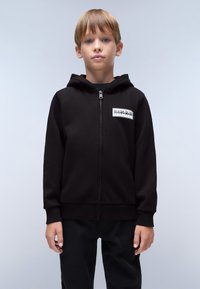 Zwarte ritssluiting hoodie van zacht materiaal, met een logo patch aan de voorkant en geribbelde manchetten. Model draagt zwarte broek.