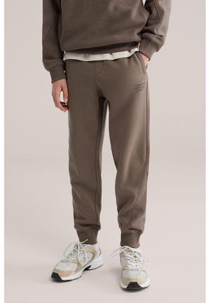 Osoba nosící hnědé teplákové kalhoty s bočními síťovanými panely a bílé tenisky, stojící s jednou rukou v kapse na jednobarevném pozadí.